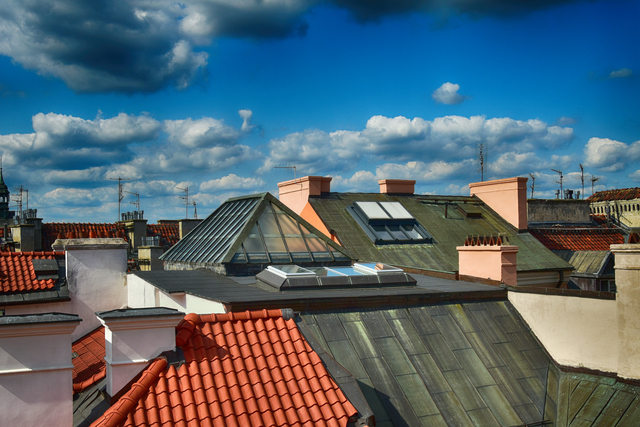 Rooftops with red clay tiles and metal skylights under a bright blue sky with scattered clouds.