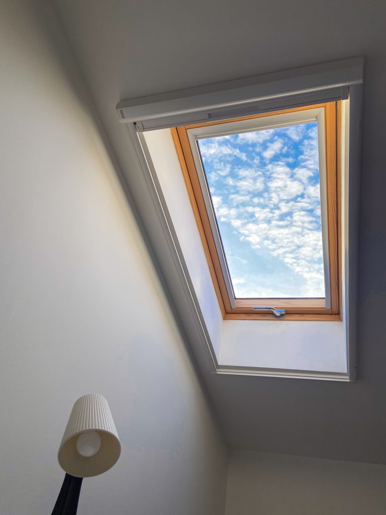 Skylight window in a sloped ceiling showing blue sky with white clouds and a wooden frame, with a floor lamp in the foreground.